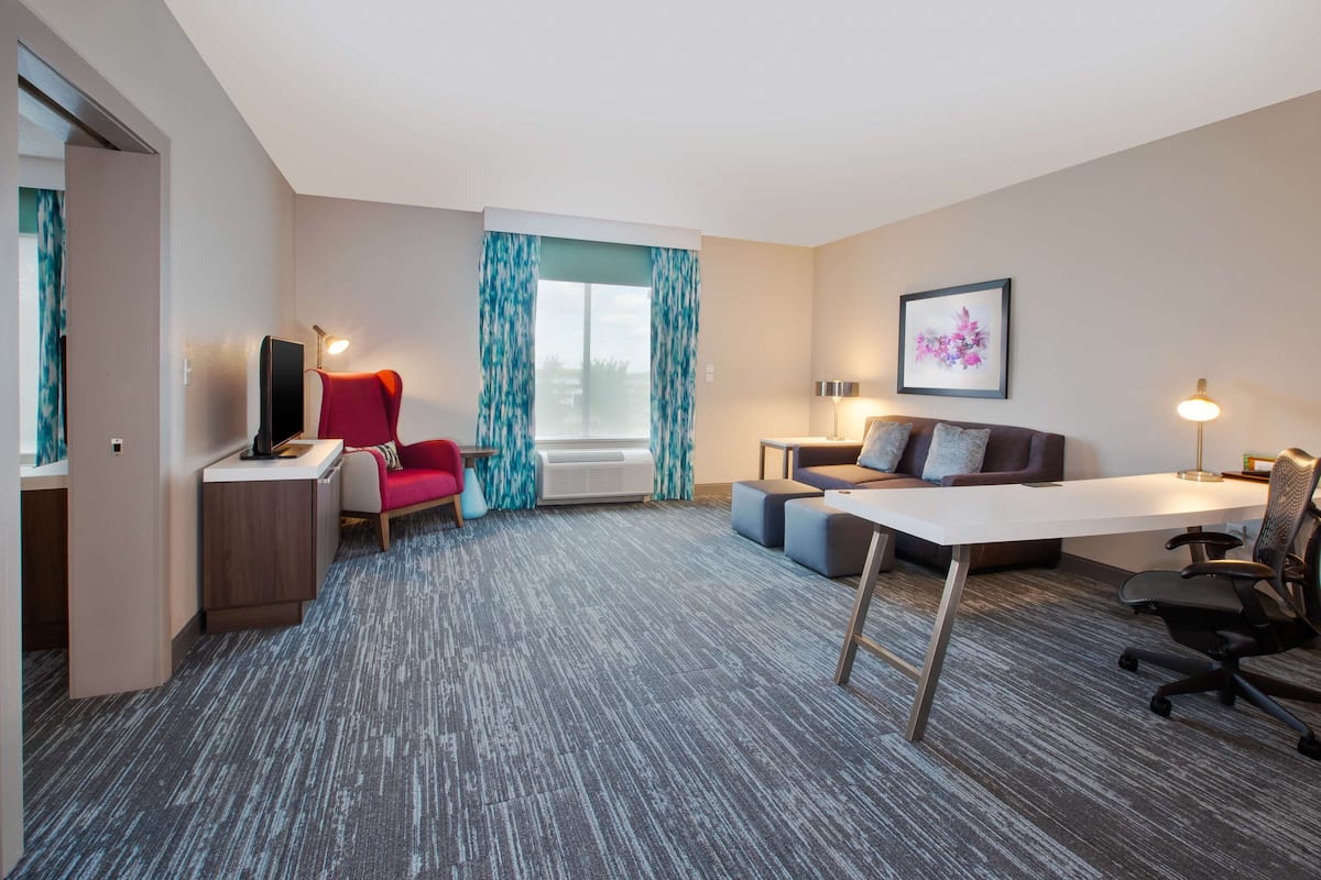 Een moderne hotelkamer met een bureau, een rode stoel, een bank en een groot raam met gordijnen.