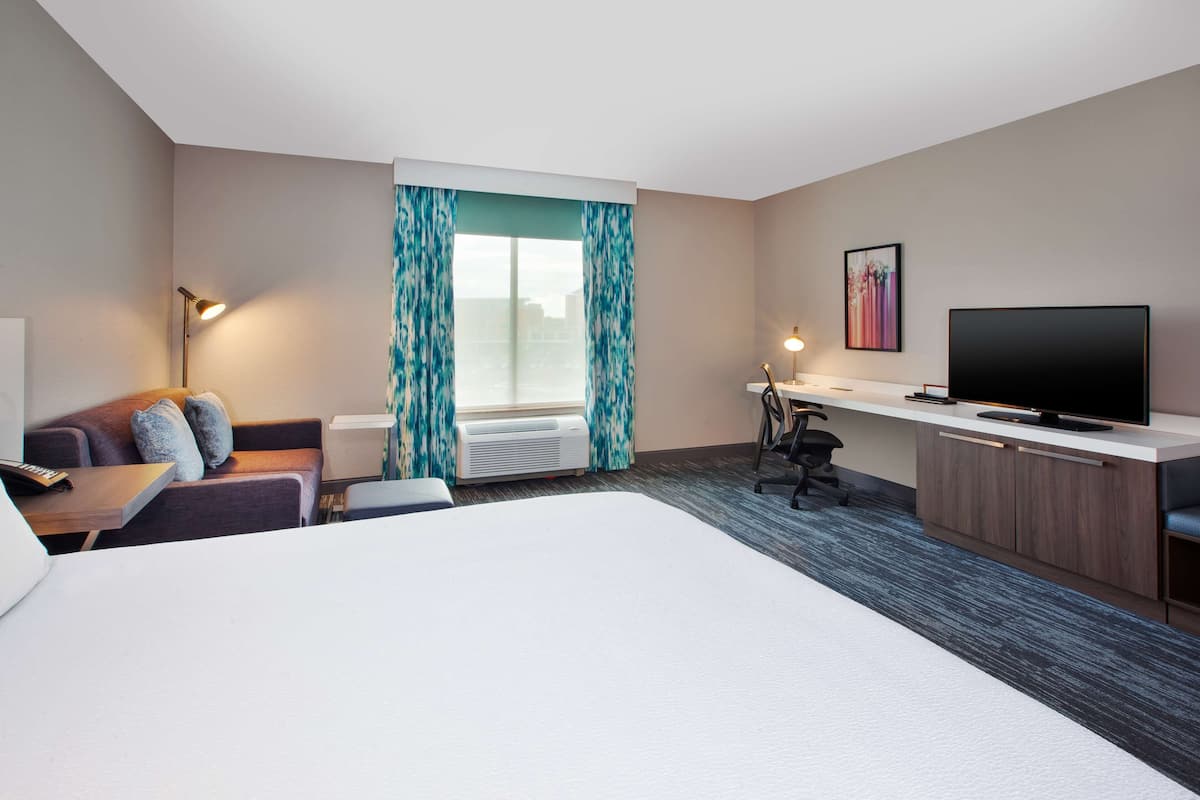 Hotelkamer met een groot bed, bureau met flatscreen televisie, sofa met kussens, staande lamp en raam met gordijnen.