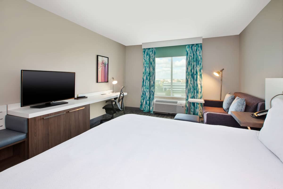 Een moderne hotelkamer met een groot bed, een flatscreen televisie, een bureau met stoel, een bank en een raam met gordijnen.