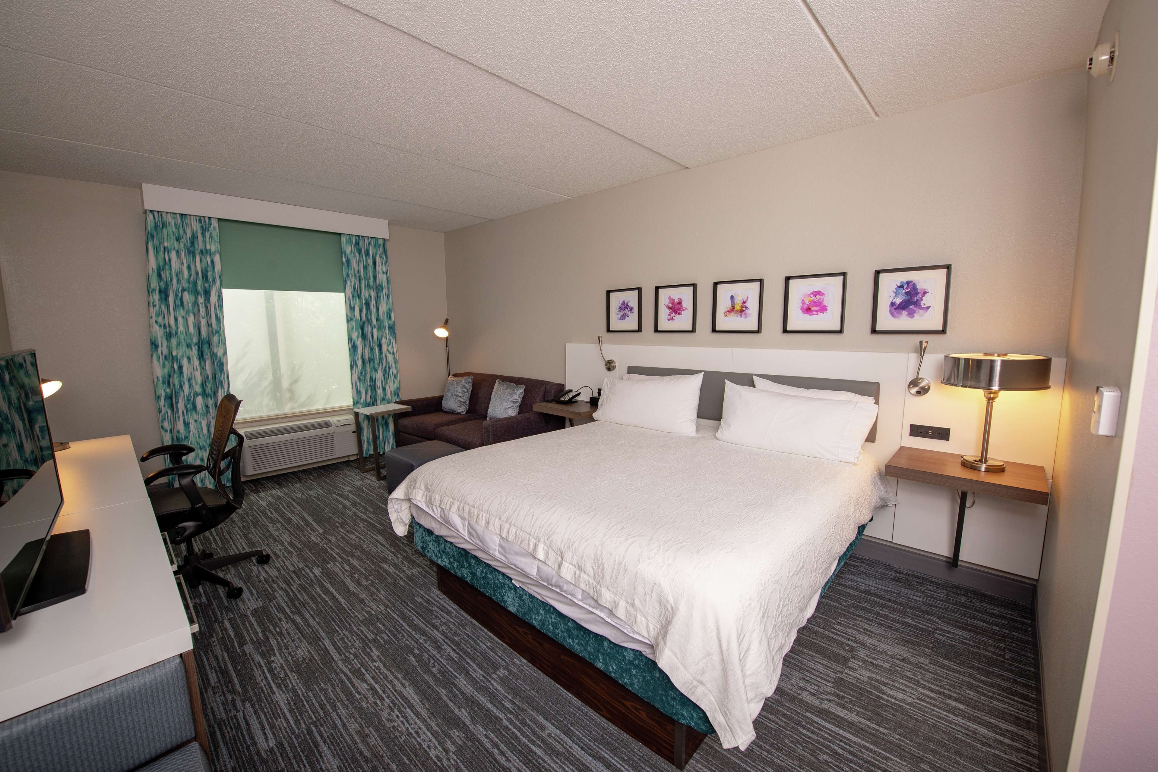 A hotel room with a large bed, a desk with a laptop, a chair, a sofa, and a window with curtains.