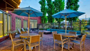 Un patio al aire libre con suelo de ladrillo hexagonal, mesas y sillas de madera y sombrillas azules.