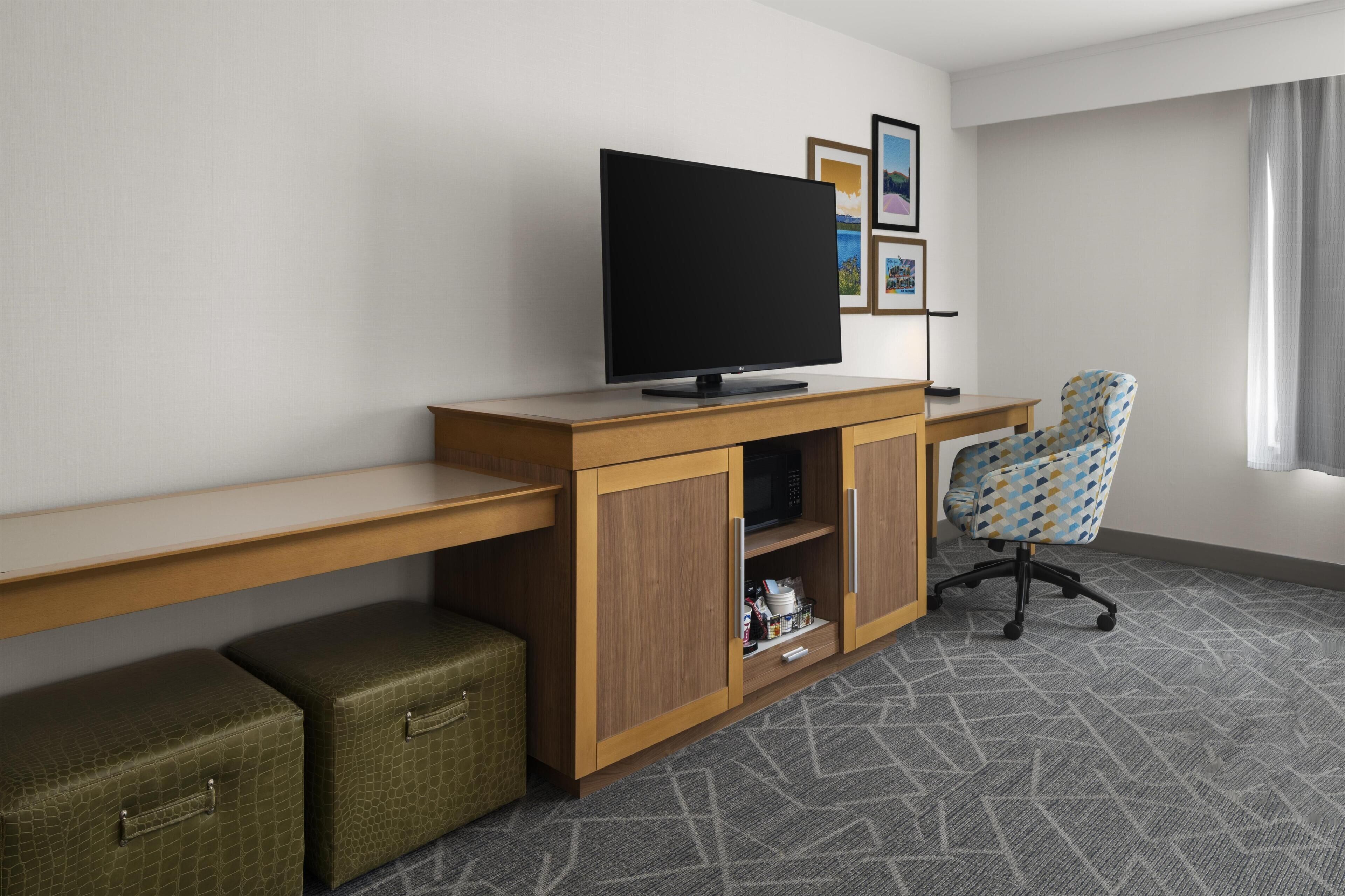 Une chambre d’hôtel avec un bureau en bois, une télévision, un fauteuil de bureau à motifs, un petit meuble et une fenêtre avec des rideaux.