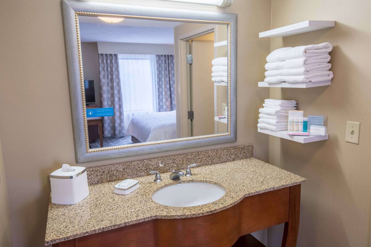 A hotel bathroom with a large mirror, a granite countertop, a sink, and a shelf with folded towels.
