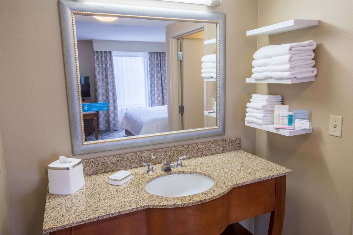 Un baño de hotel con un espejo grande, encimera de granito, lavamanos y un estante con toallas dobladas.