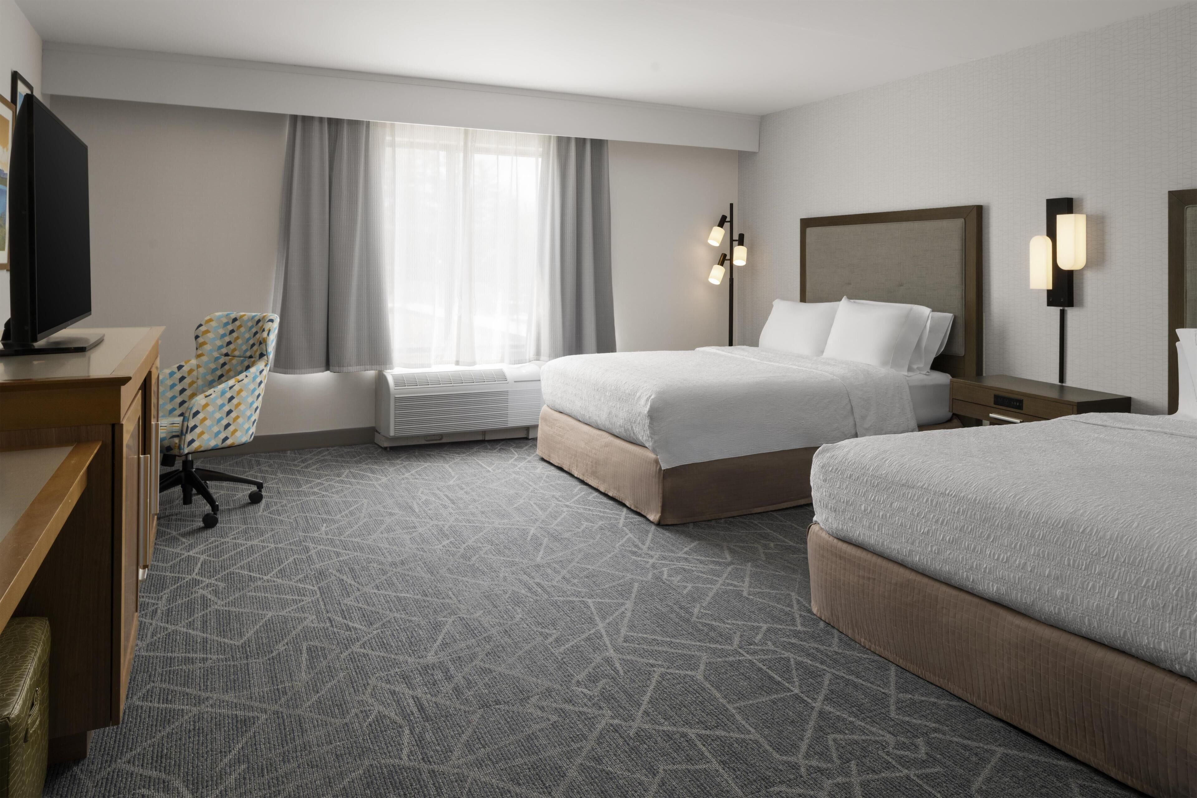 Habitación de hotel con dos camas, un escritorio, una silla, un televisor, una ventana con cortinas y una lámpara de pared.