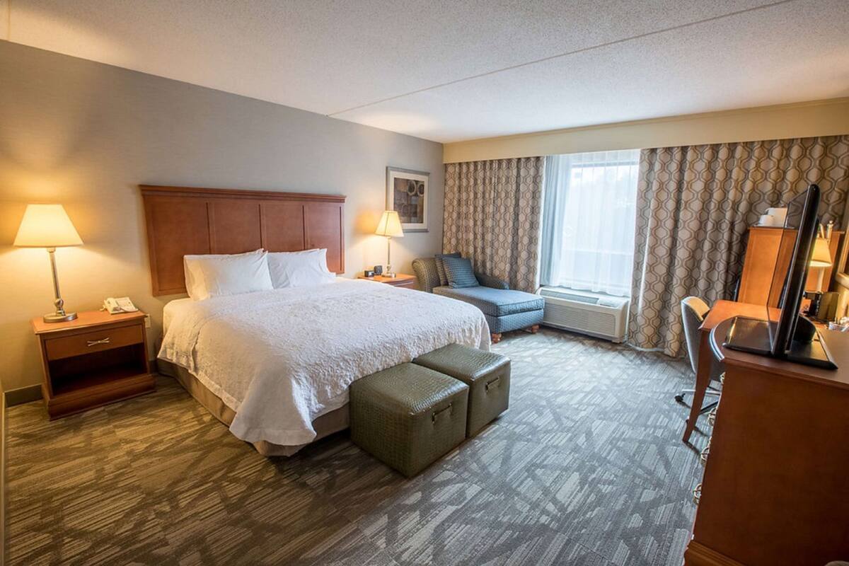 A hotel room with a bed, bedside tables, a desk, a chair, a window with curtains, and a wall-mounted TV.