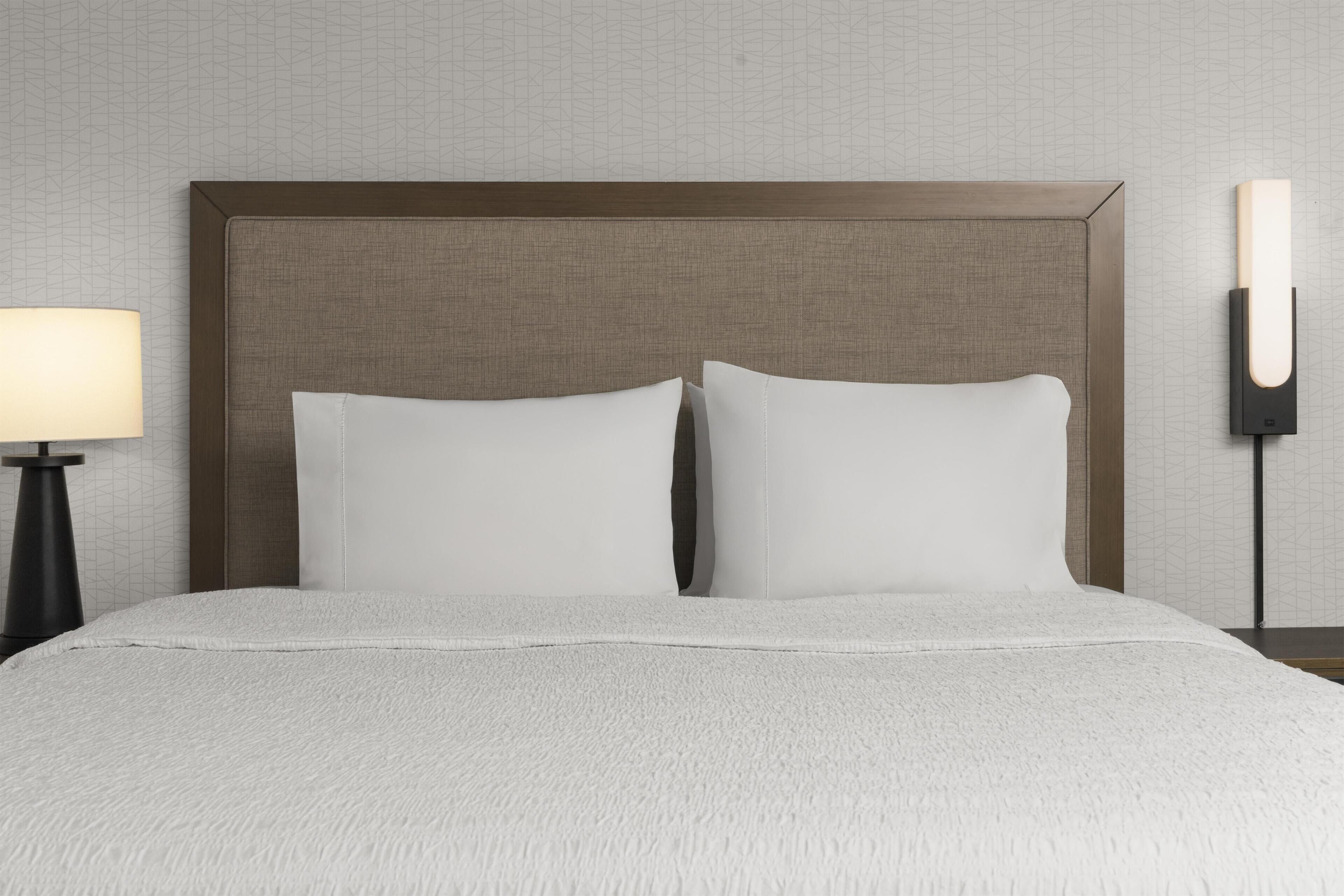 Una cama con cabecera, dos almohadas, una lámpara de noche y una lámpara de pared.