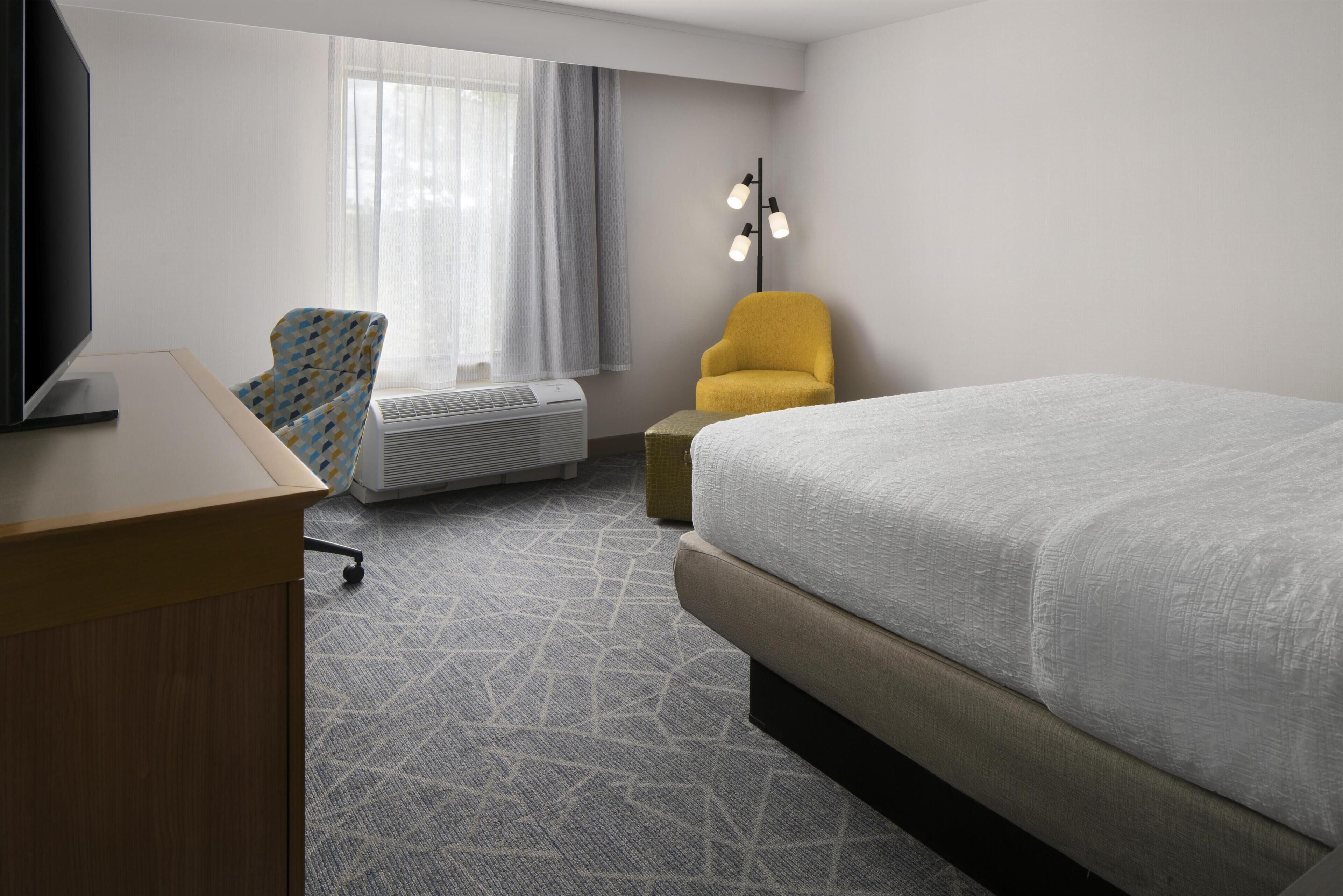 A hotel room with a bed, a desk with a flat-screen TV, a chair, an air conditioning unit, and a floor lamp.