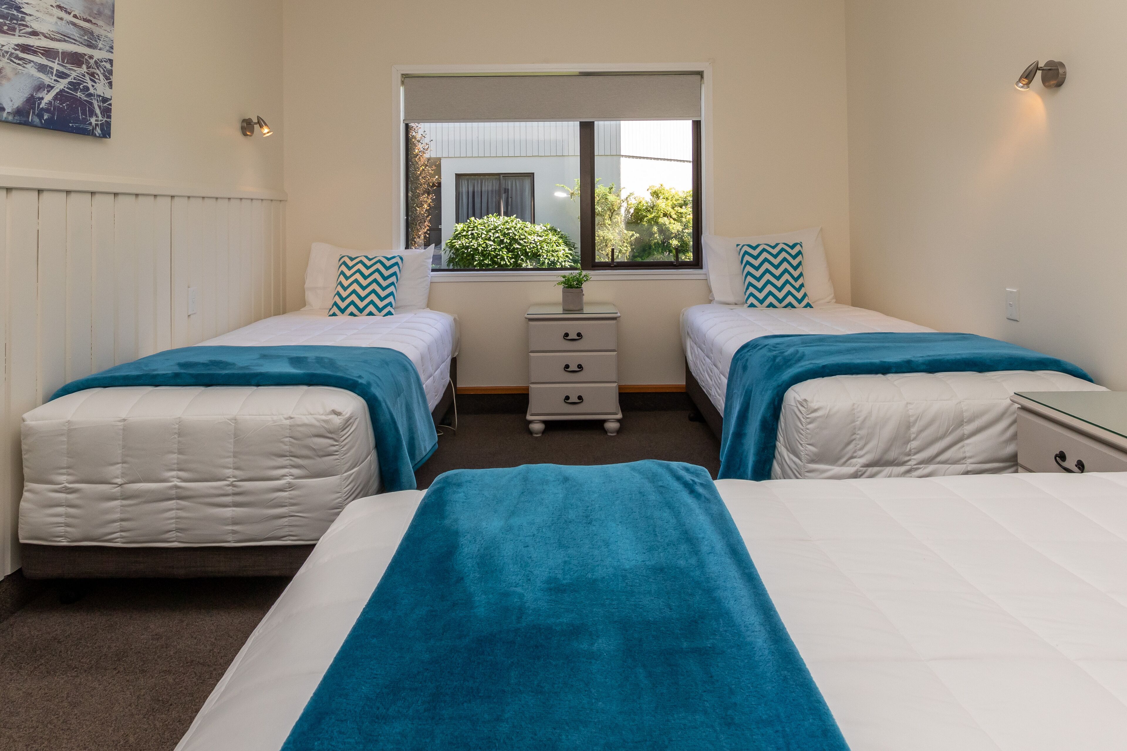 A twin room with two beds, a small bedside table, a window with a view of greenery, and a wall-mounted light fixture.