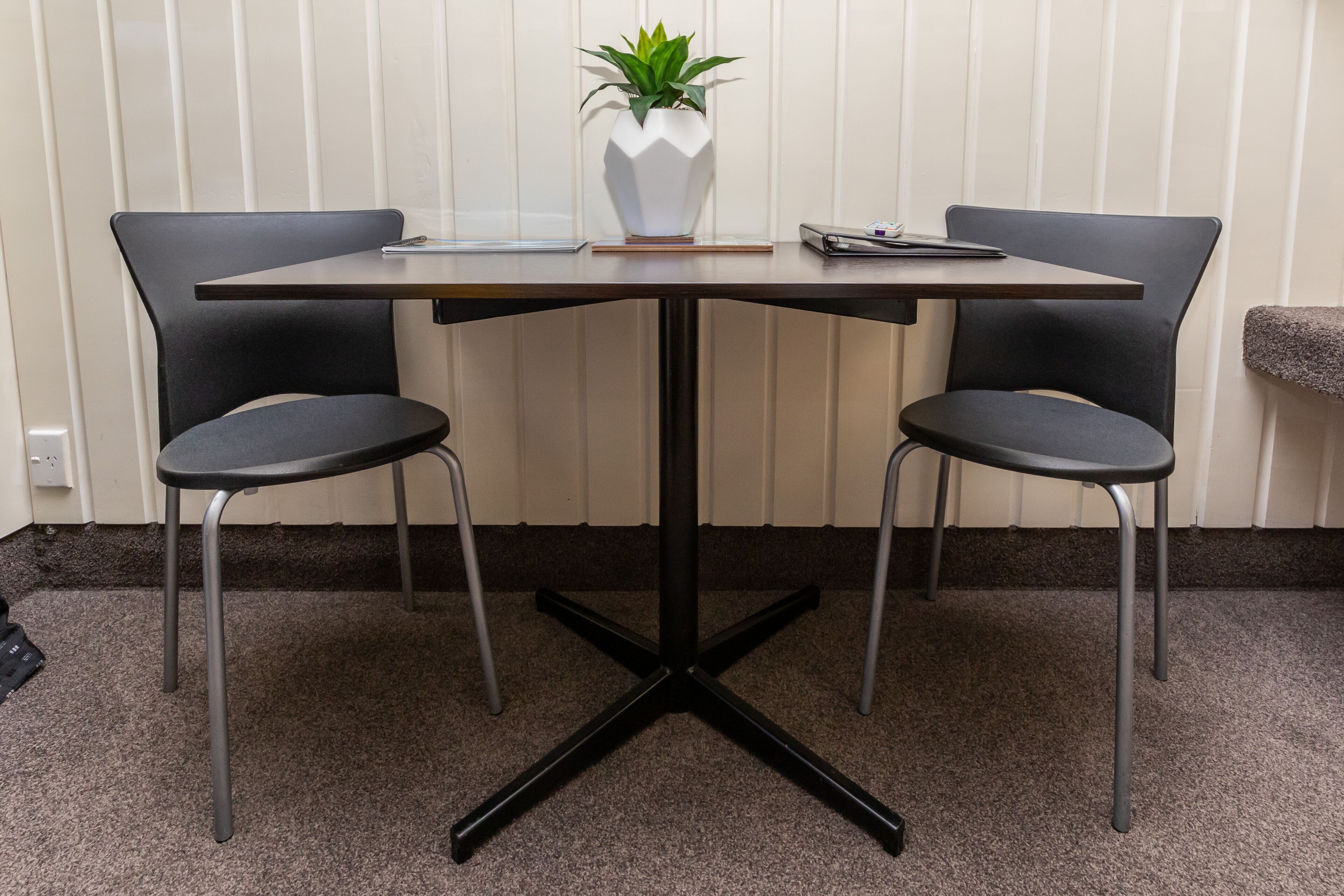 Una pequeña mesa de reuniones con dos sillas, un jarrón blanco con una planta y una tableta sobre la mesa.