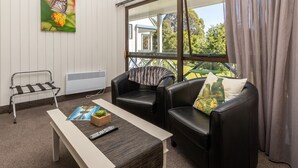 A room with a white wall, a large window with curtains, two black leather sofas, a small table with a remote control, and a picture of a butterfly on the wall. - ASURE Explorer Motel & Apartments (Te Anau)
