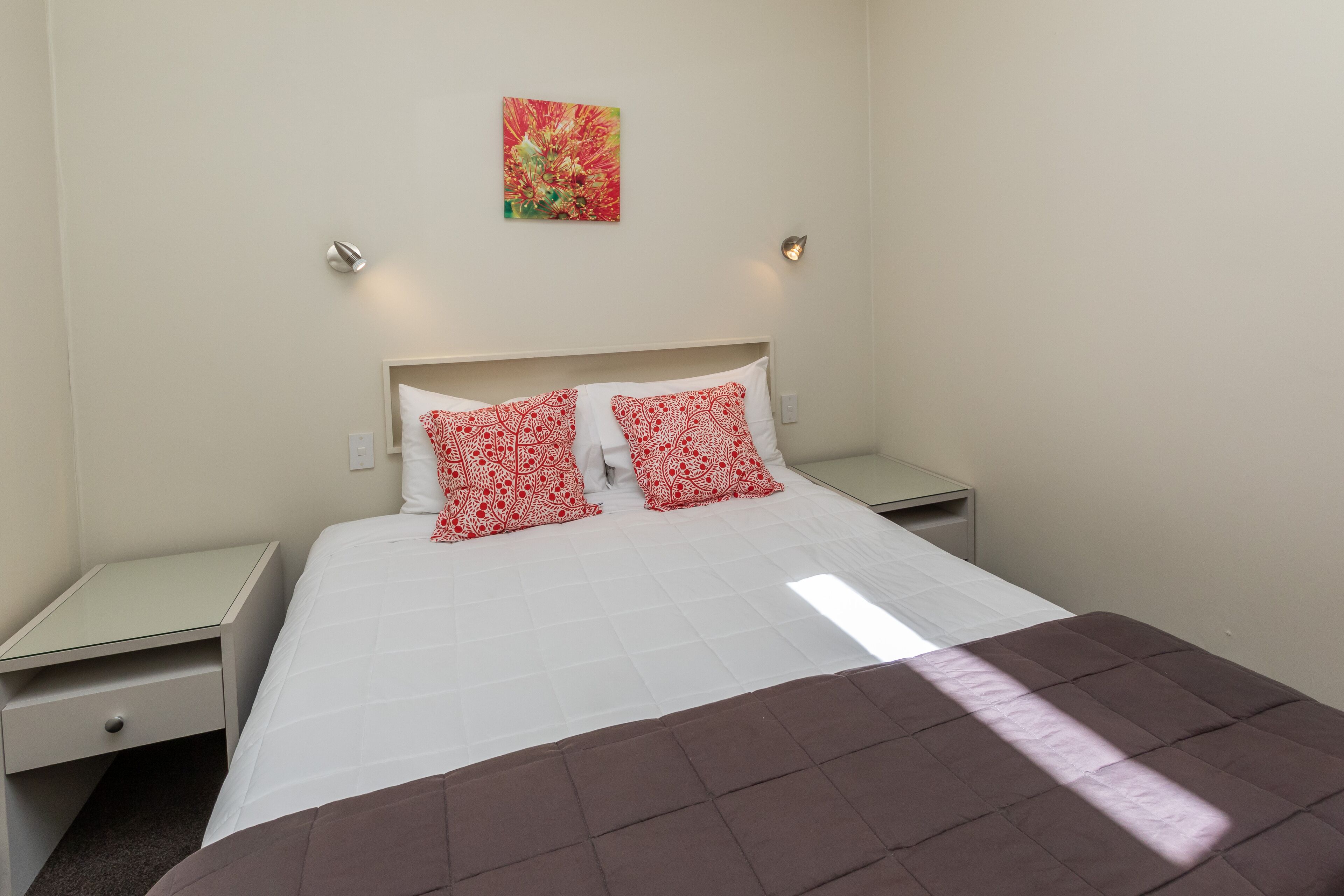 A hotel room with a bed, two bedside tables, a nightstand, a wall-mounted light, and a painting.