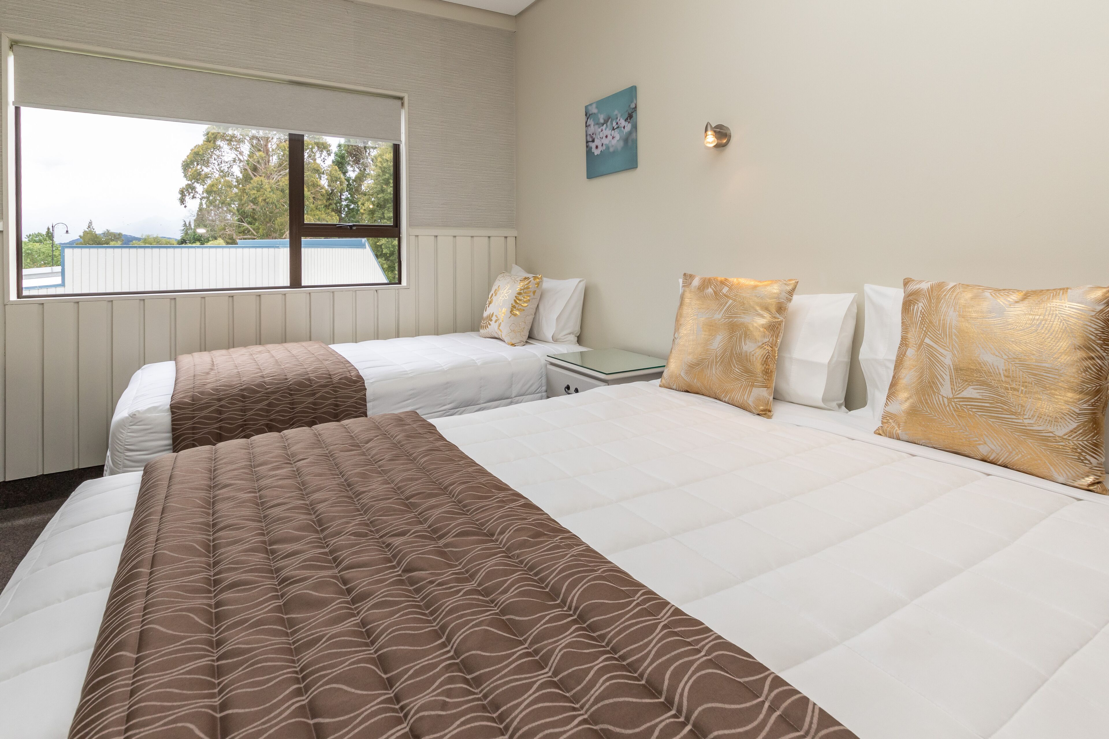 A hotel room with two beds, a window with a view, a small table, and a wall-mounted light.