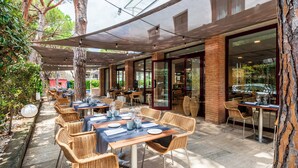 Un espacio de comedor al aire libre con sillas y mesas de mimbre, ventanas amplias y una pared de ladrillo.
