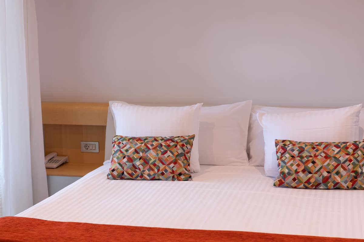A hotel room with a bed, two patterned pillows, a white blanket, a wooden headboard, a nightstand with a telephone, and a power outlet.