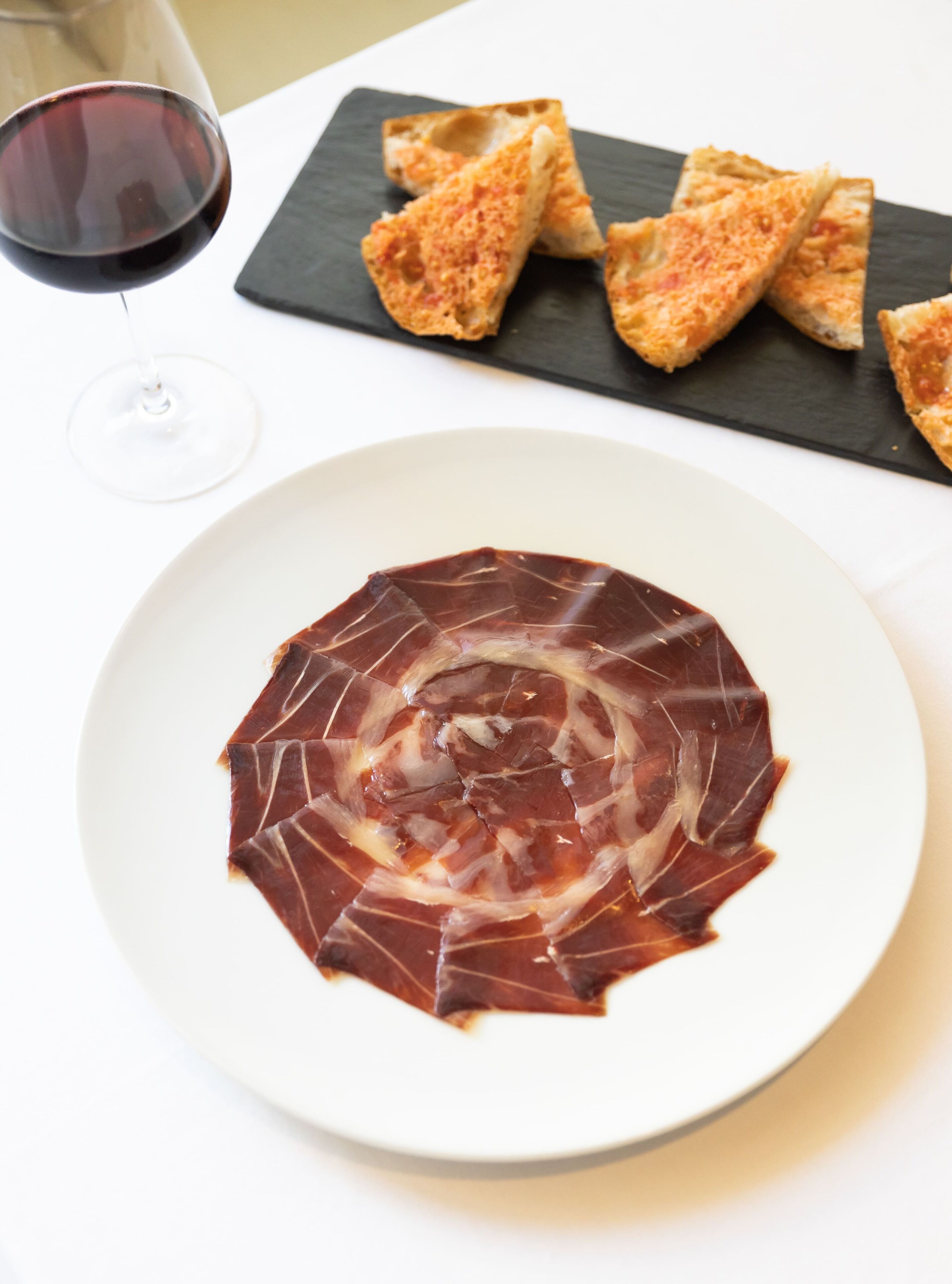 Um prato com fatias finas de carne curada dispostas em espiral, acompanhado por um copo de vinho tinto e uma lousa de ardósia com pedaços de pão.