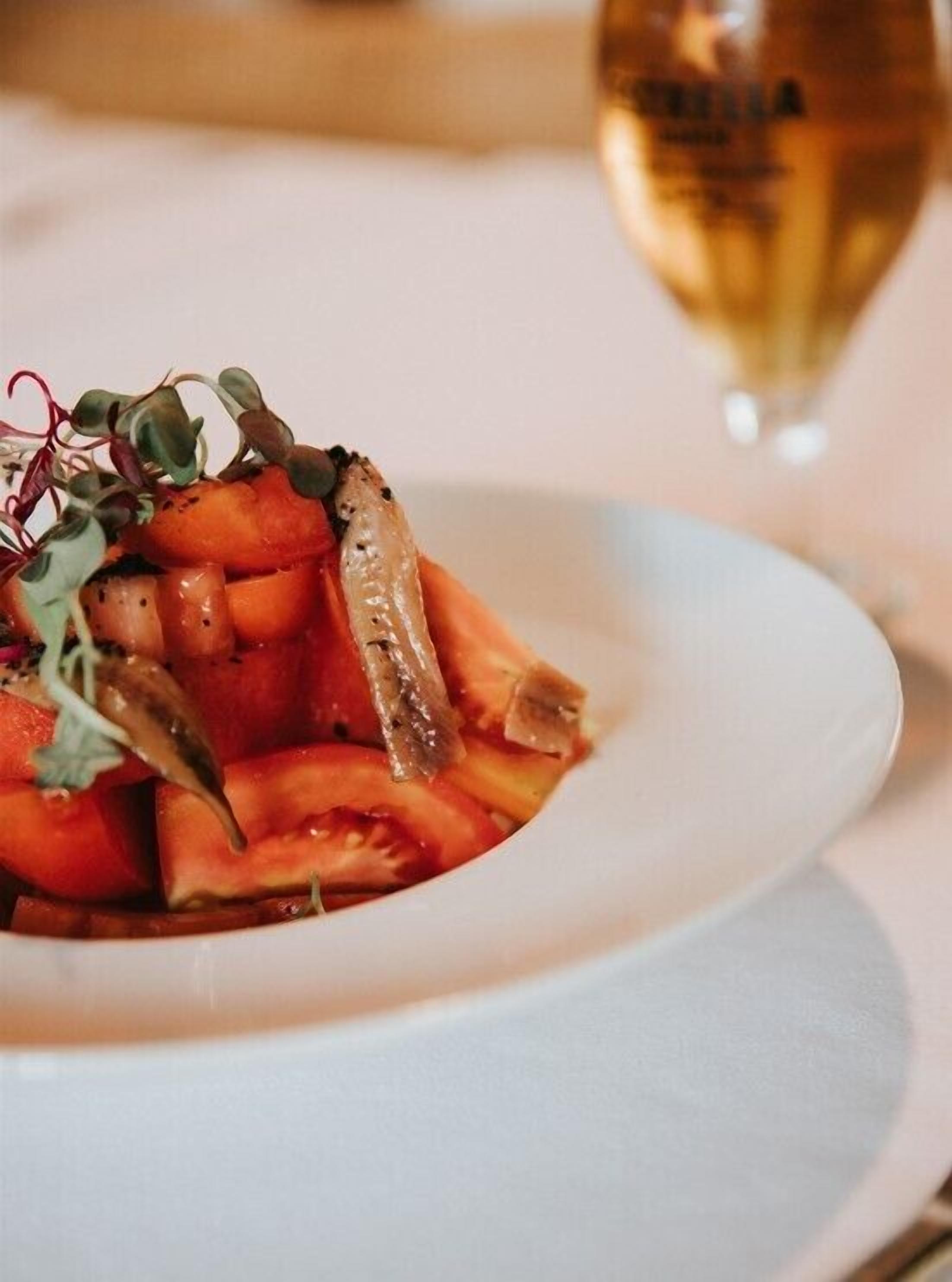A white plate with a dish of sliced tomatoes and mushrooms, garnished with microgreens, on a table with a glass of white wine in the background.