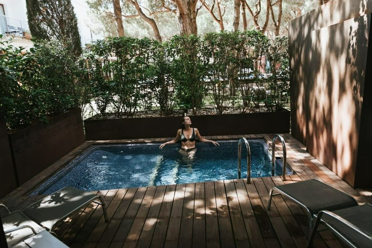 A person in a swimming pool with wooden deck chairs and a metal ladder.