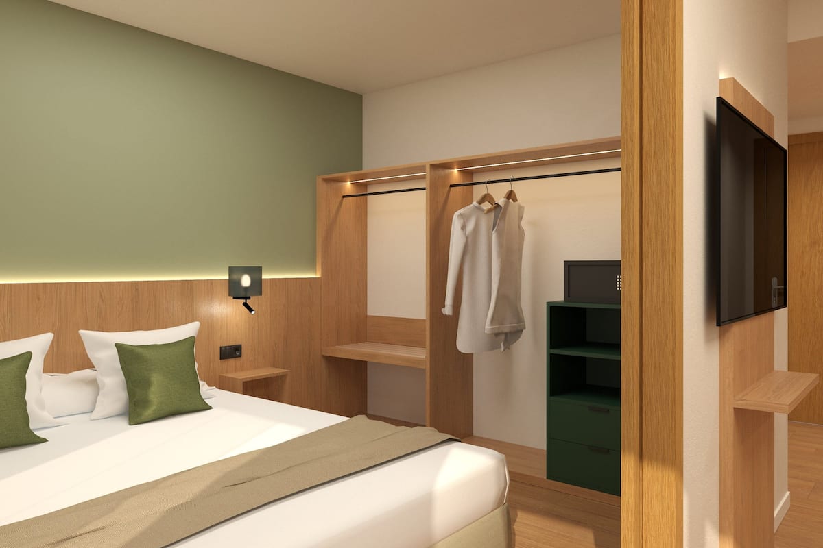 A modern hotel room with a bed, green pillows, a bedside table, a TV, a wardrobe, and a green storage unit.