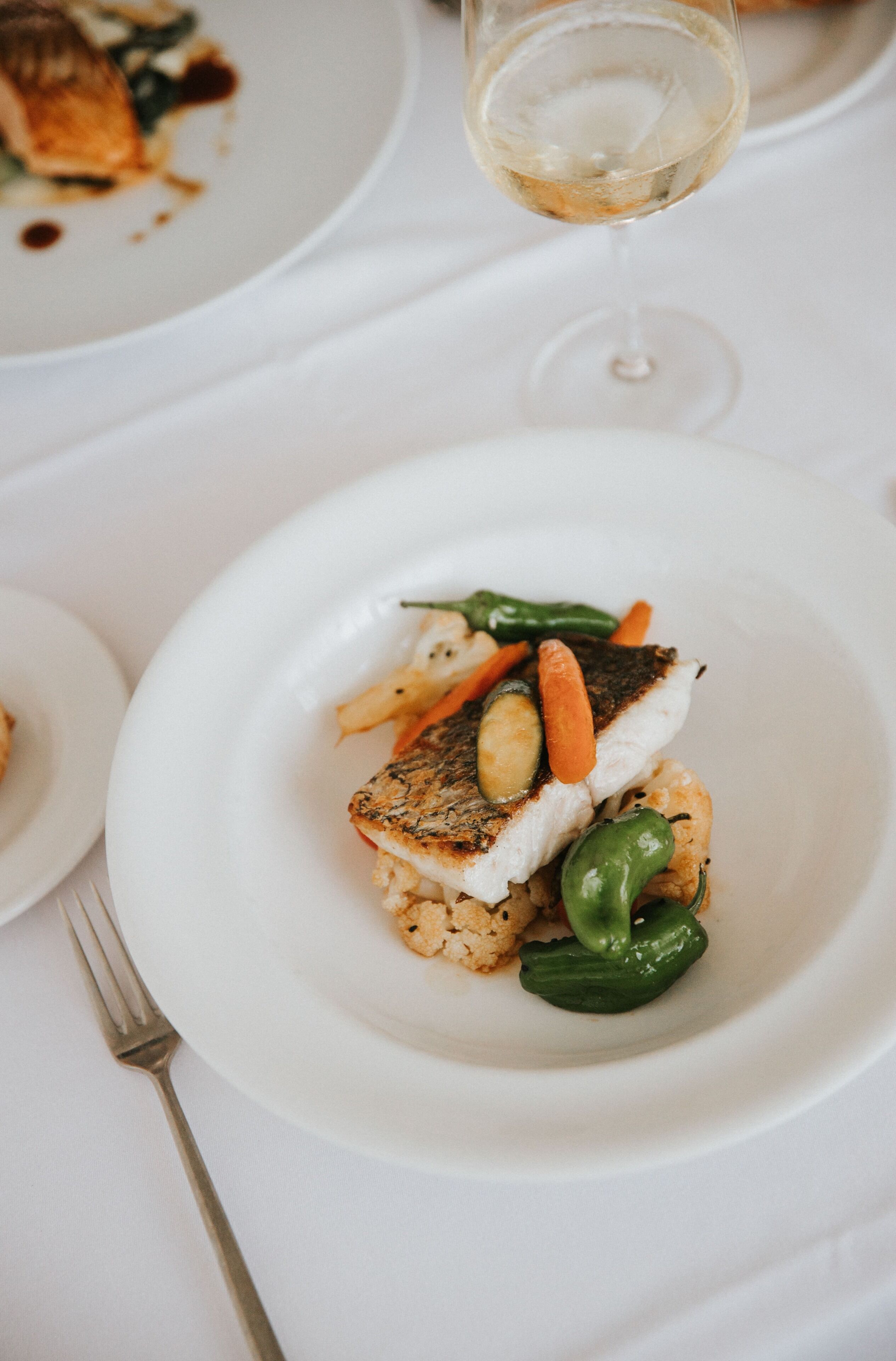 A white plate with a piece of fish, vegetables, and a glass of white wine on a white tablecloth.