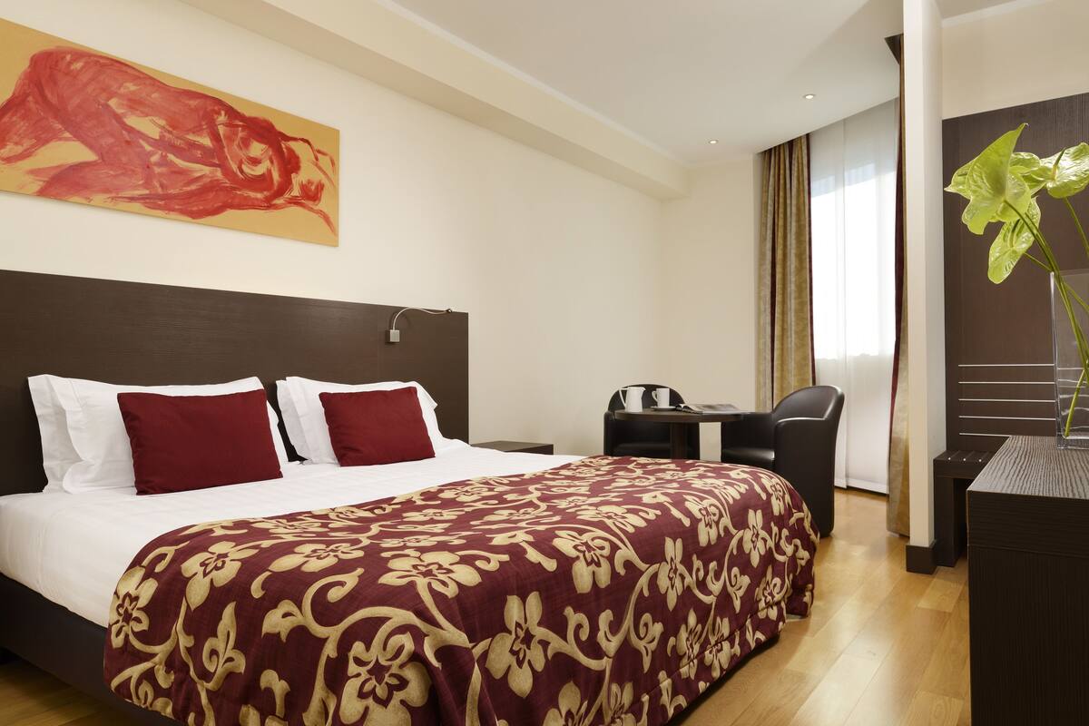 A hotel room with a large bed, a desk, a chair, and a plant.