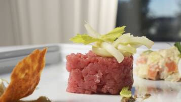 Un plat composé de viande crue finement hachée, peut-être du thon, agrémenté de feuilles vertes, servi dans une assiette blanche avec une portion de légumes marinés et un cracker croustillant.