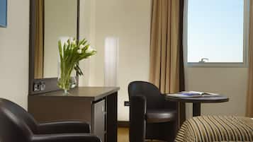 Hotelkamer met bureau, twee stoelen, een gestreepte bank, een spiegel, een vaas met bloemen en een raam met gordijnen.