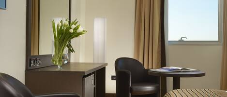 A hotel room with a desk, two chairs, a striped sofa, a mirror, a vase with flowers, and a window with curtains.