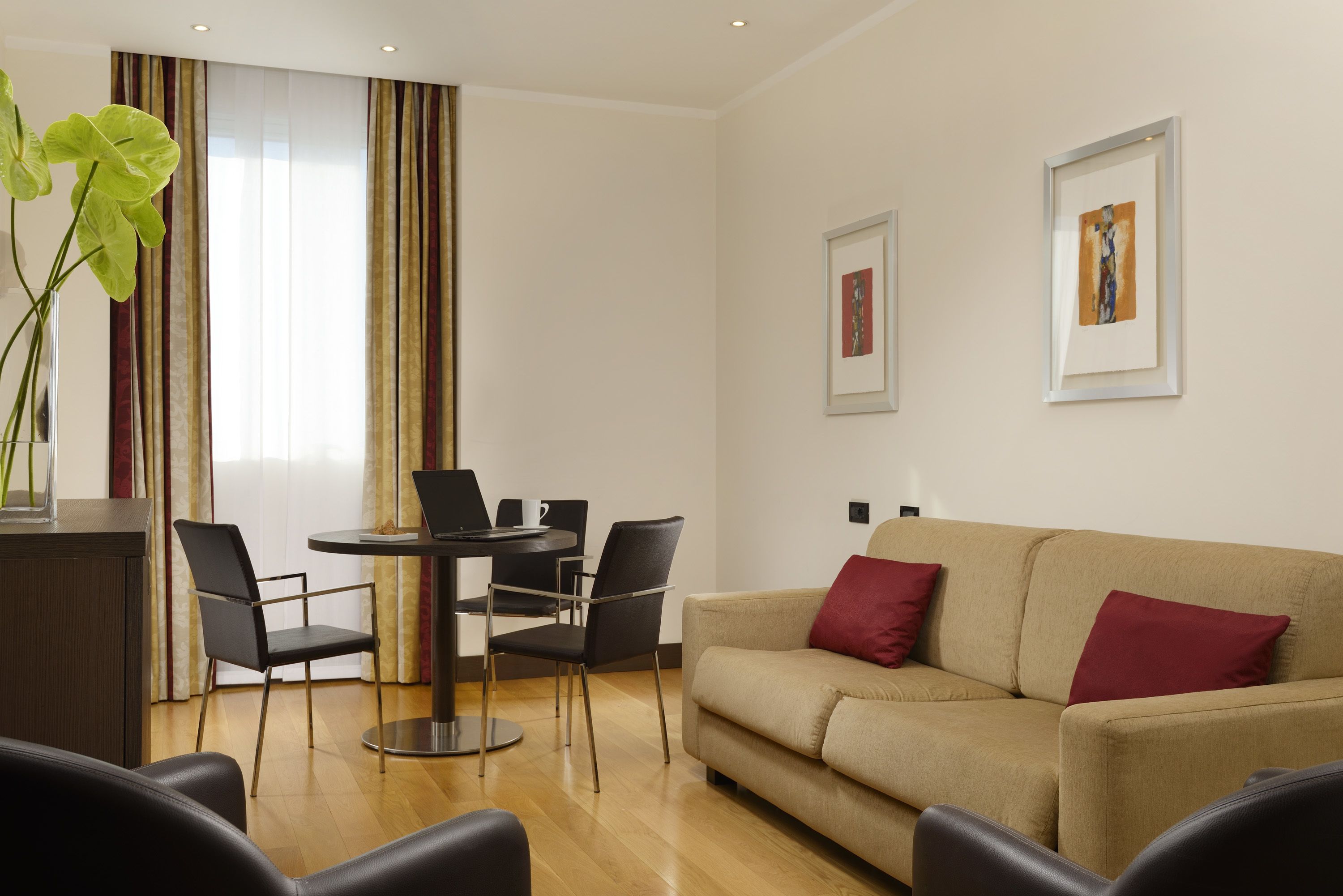 A modern living room with a beige sofa, two red cushions, a dining table with four chairs, and two framed artworks on the wall.