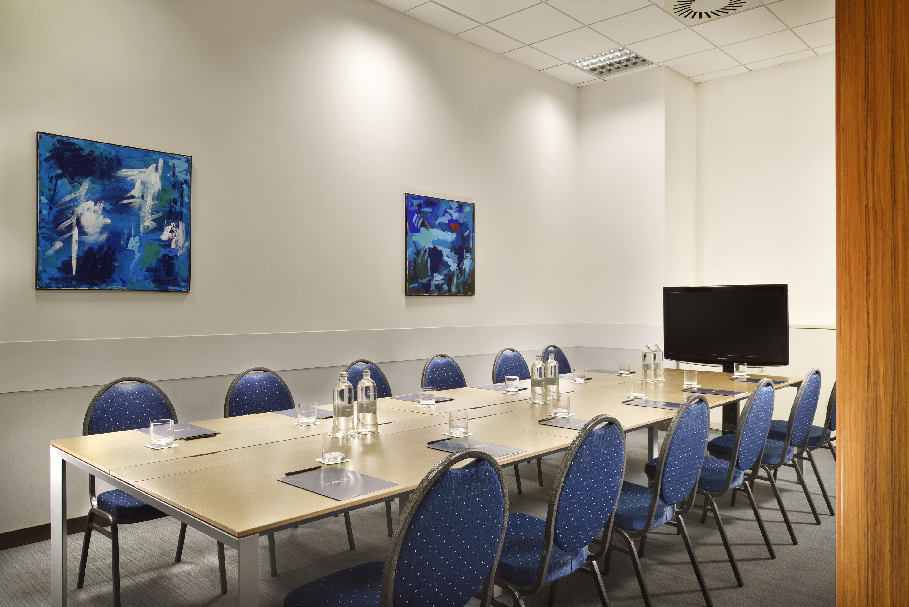 Una sala de conferències amb una taula llarga, cadires blaves, un televisor de pantalla plana i dos quadres abstractes a les parets.