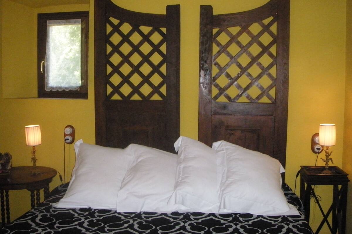 Un lit avec des oreillers blancs et une couvre-lit à motifs noir et blanc, dans une pièce aux murs jaunes. On y trouve deux têtes de lit en bois ornées de motifs en treillis, ainsi que deux lampes de chevet posées sur des tables de chevet en bois.