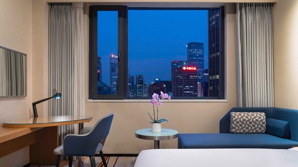 Quarto de hotel com escrivaninha, cadeira, cama e vista para a paisagem urbana ao entardecer.