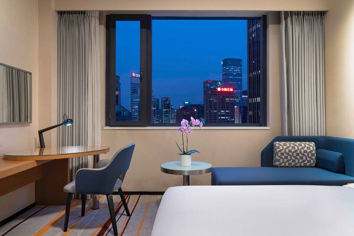 Habitación de hotel con escritorio, silla, cama y vista a un paisaje urbano al atardecer.
