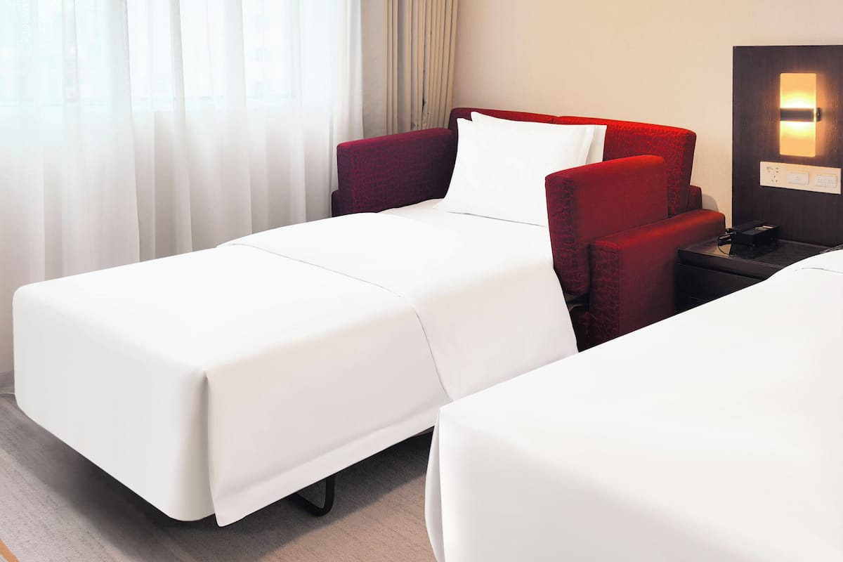 Une chambre d’hôtel avec deux lits, un fauteuil rouge recouvert de tissu, une table de chevet avec une lampe et une fenêtre avec des rideaux légers.