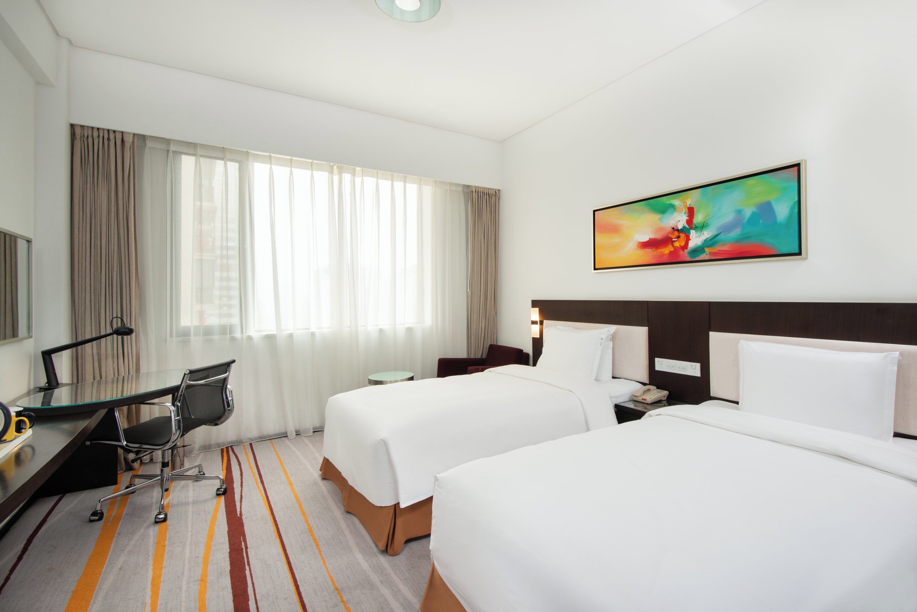 A hotel room with two beds, a desk with a chair, a telephone, and a painting on the wall.