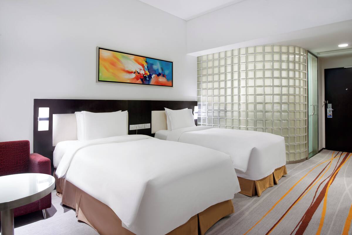 Habitación de hotel con dos camas, una silla roja, una mesita pequeña y un cuadro abstracto de colores en la pared.