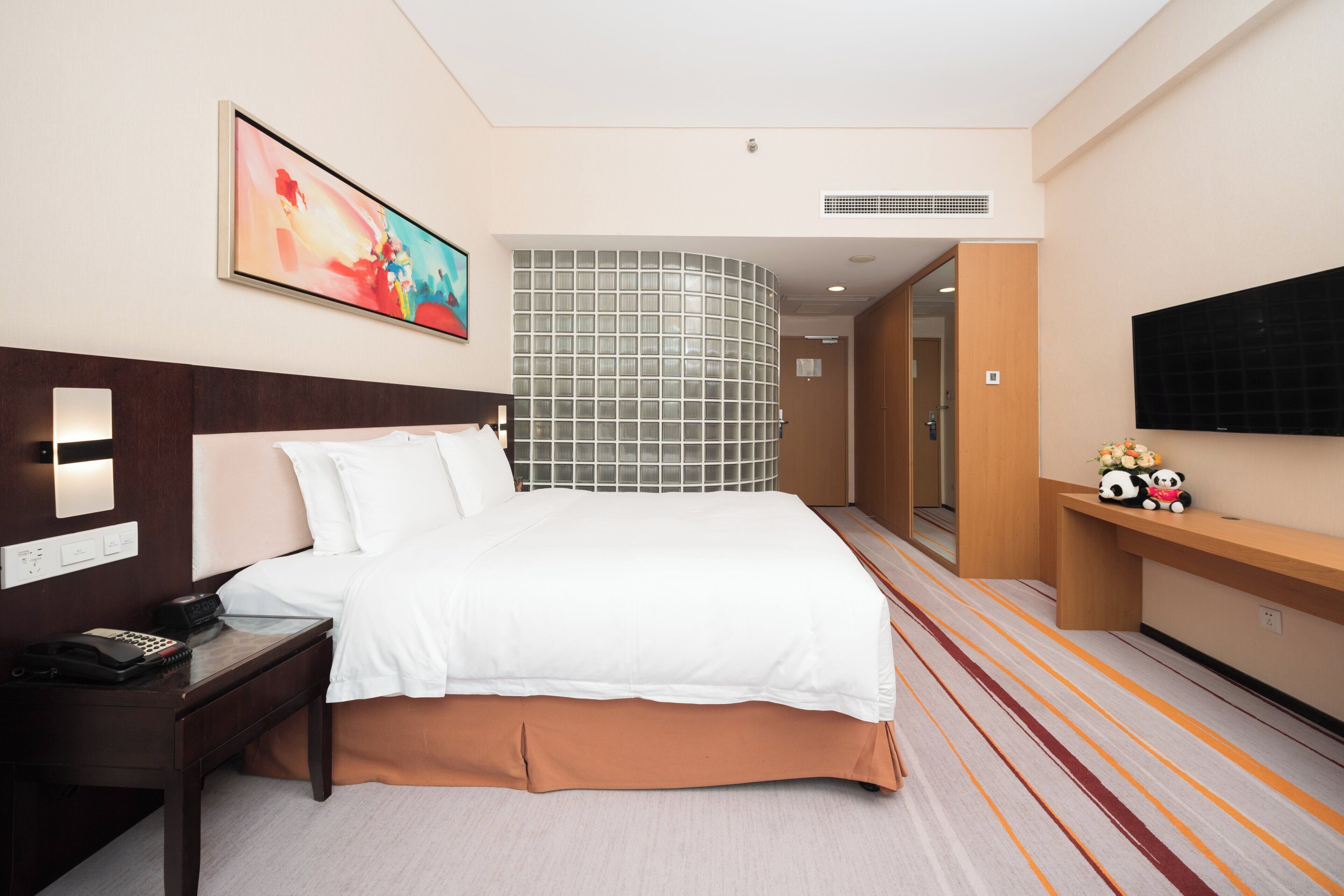 A hotel room with a bed, bedside table, a painting, a television, and a decorative shelf with plush toys.