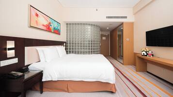 A hotel room with a bed, bedside table, a painting, a television, and a decorative shelf with plush toys.
