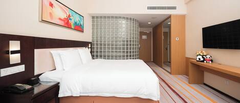 A hotel room with a bed, bedside table, a painting, a television, and a decorative shelf with plush toys.