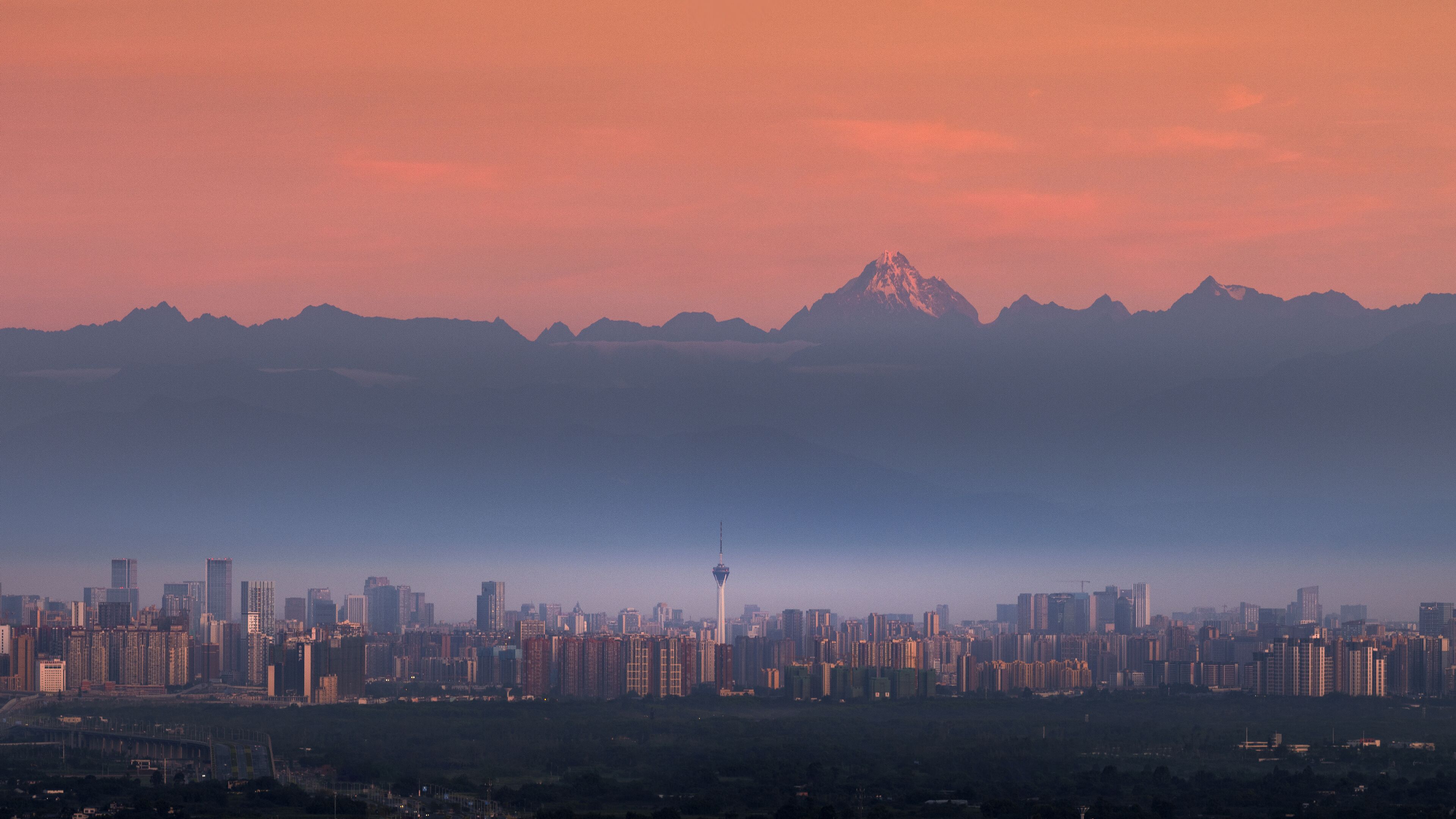 A cityscape with a prominent tower, a mountain range in the background, and dense urban buildings.