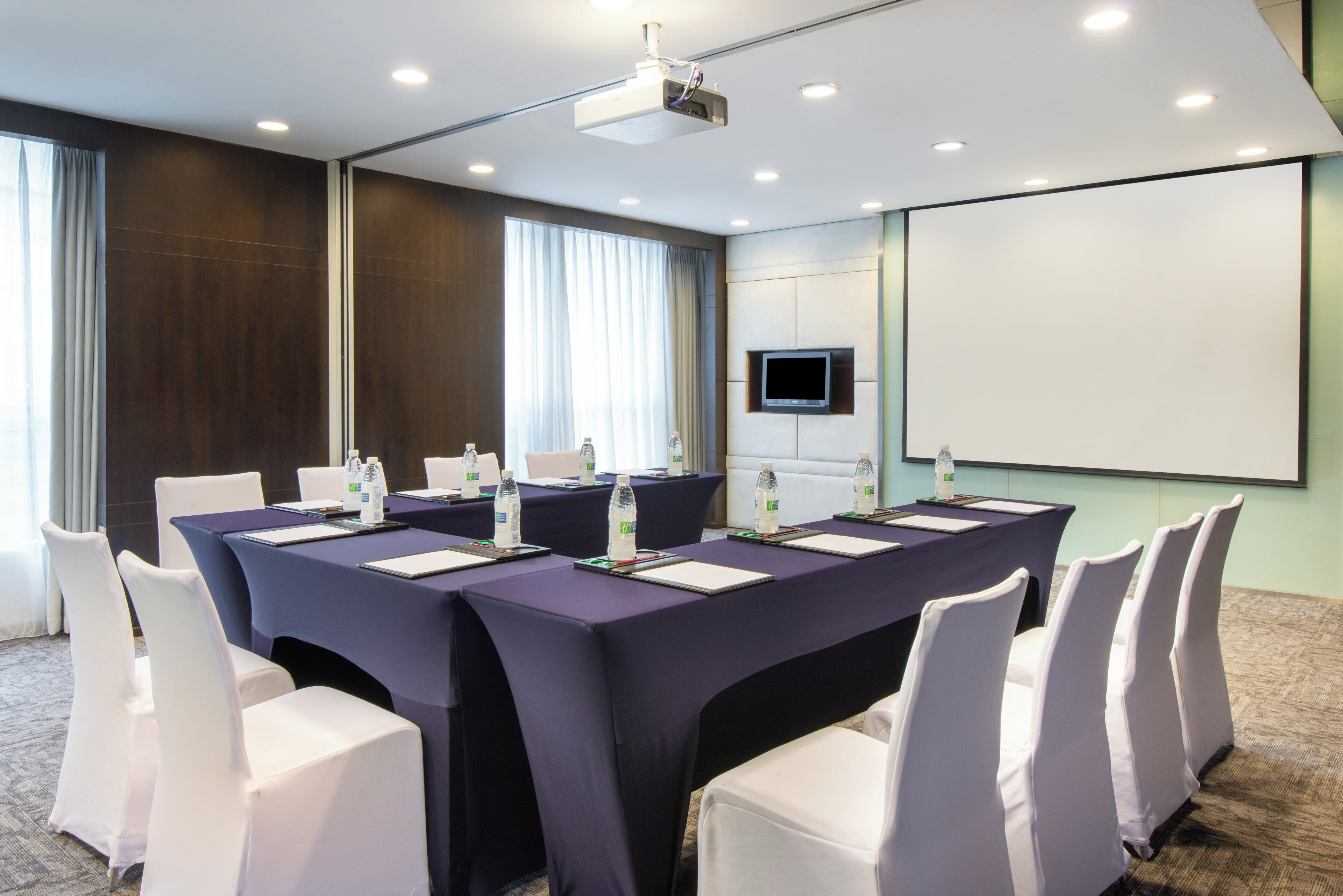 A conference room with a long table, white chairs, water bottles, and a projector screen.