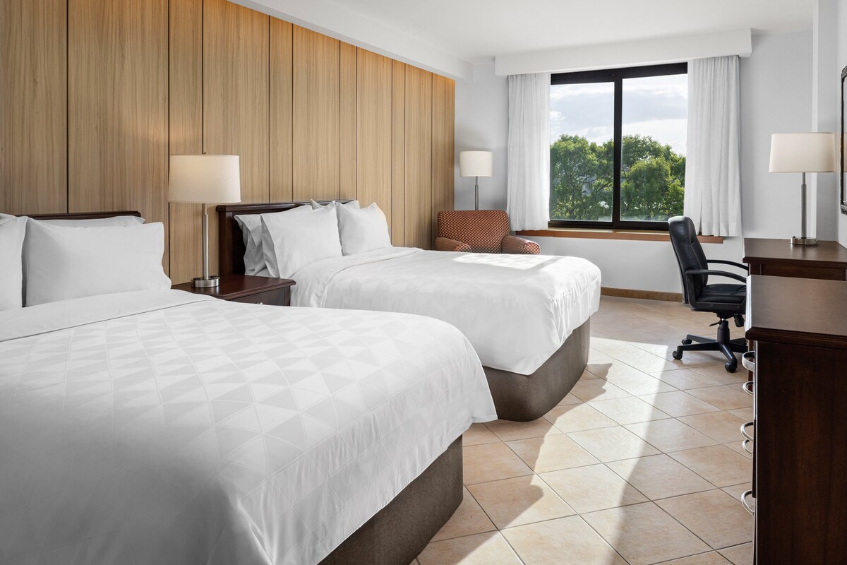 A hotel room with two beds, a desk, a chair, and a window with a view of trees.