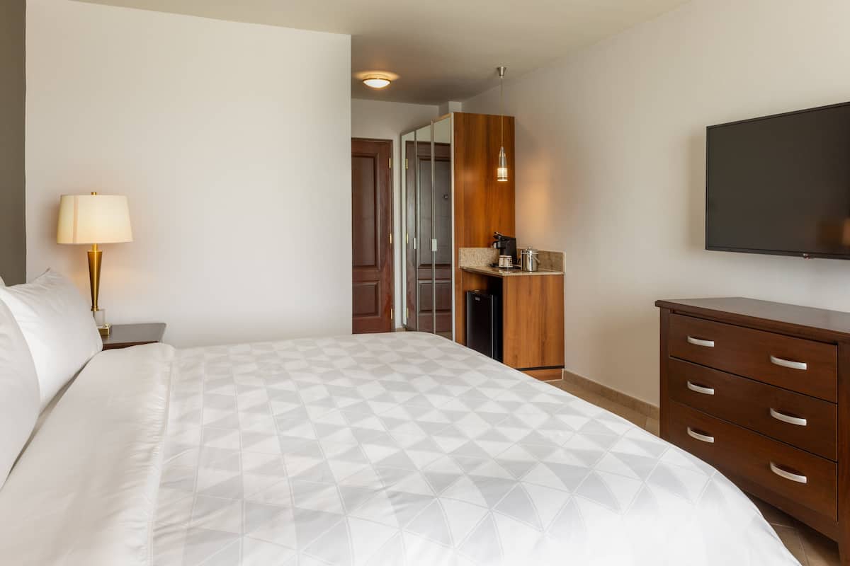 A hotel room with a bed, a nightstand, a lamp, a wooden dresser, a television, and a kitchenette area.
