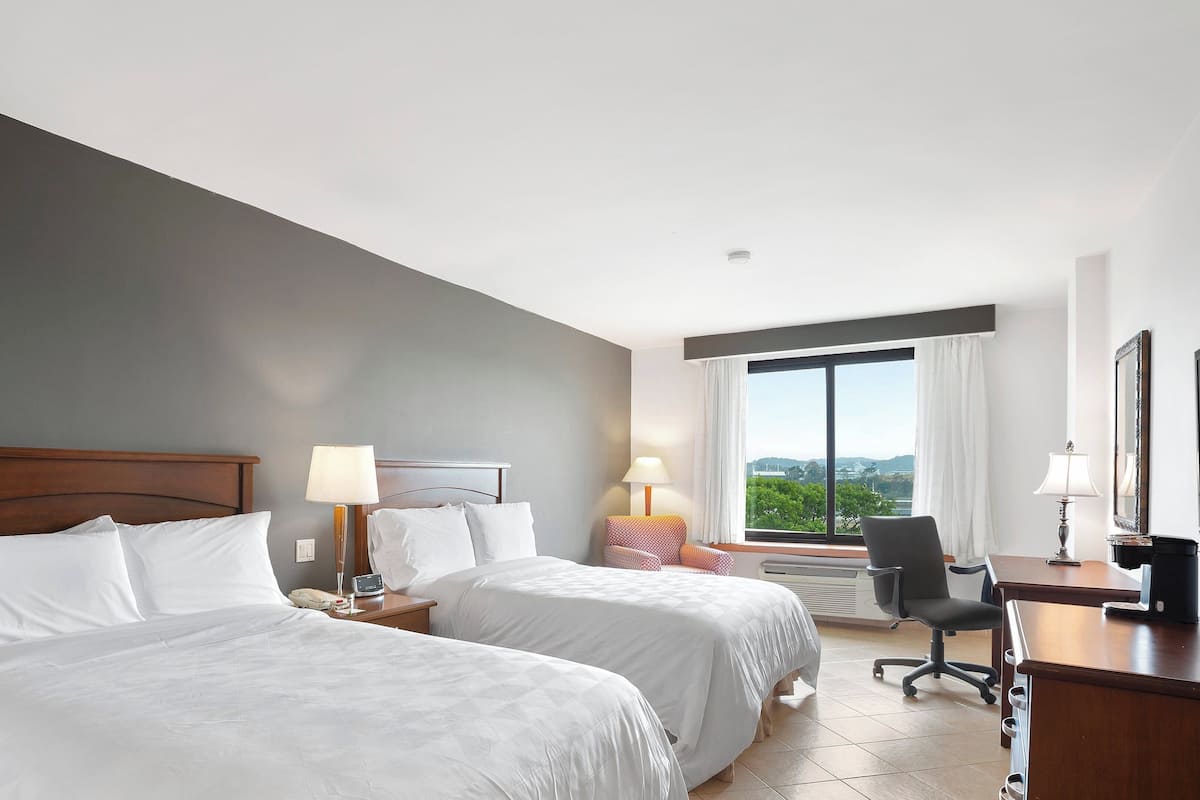 A hotel room with two beds, a desk, a chair, and a large window offering a view.