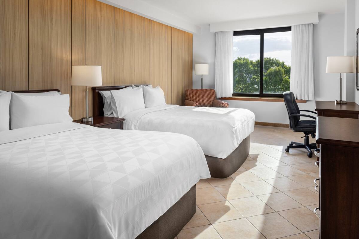 A hotel room with two beds, a desk, a chair, and a window with a view of trees.