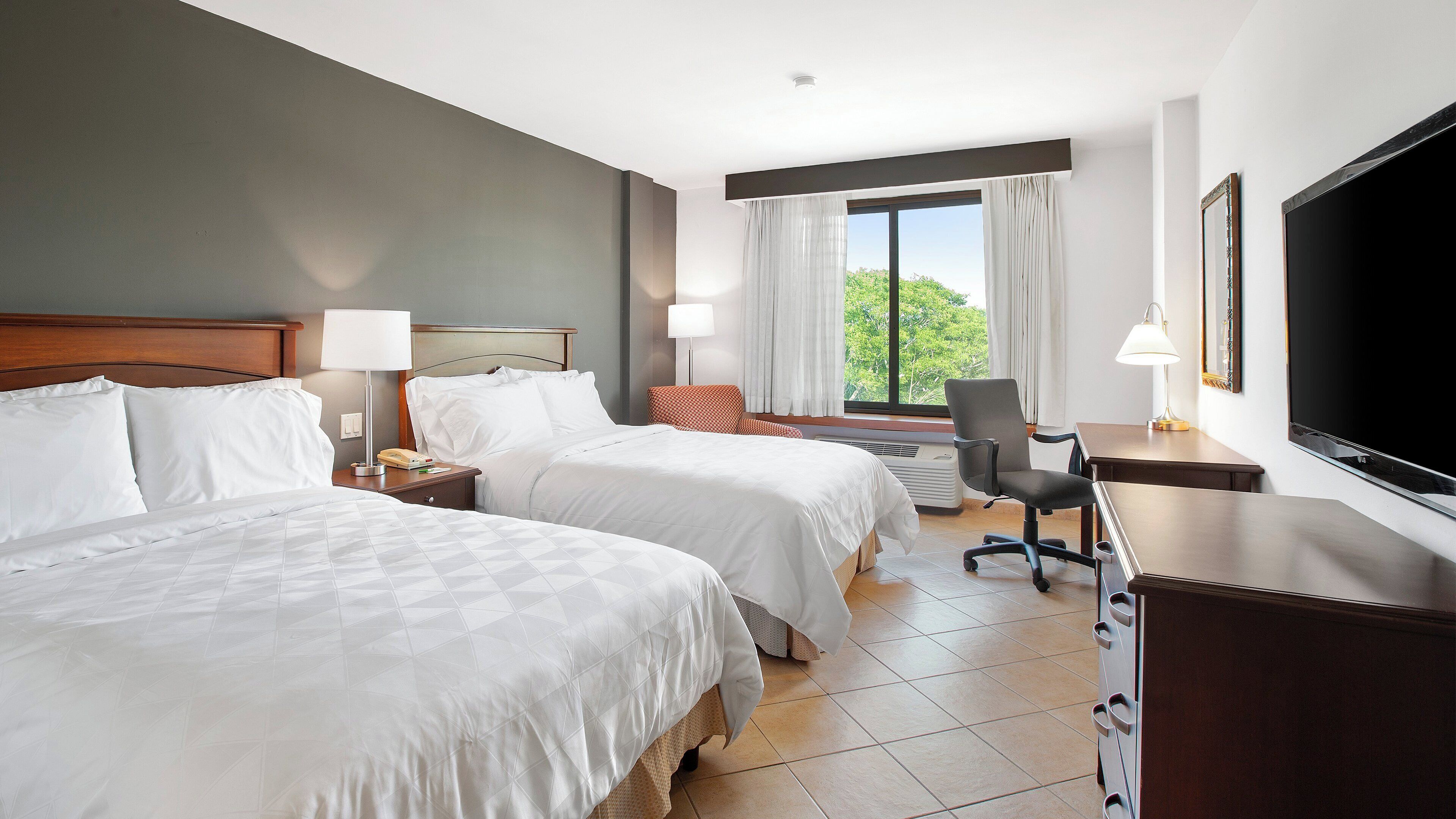 A hotel room with two beds, a desk, a chair, a television, and a window with curtains.
