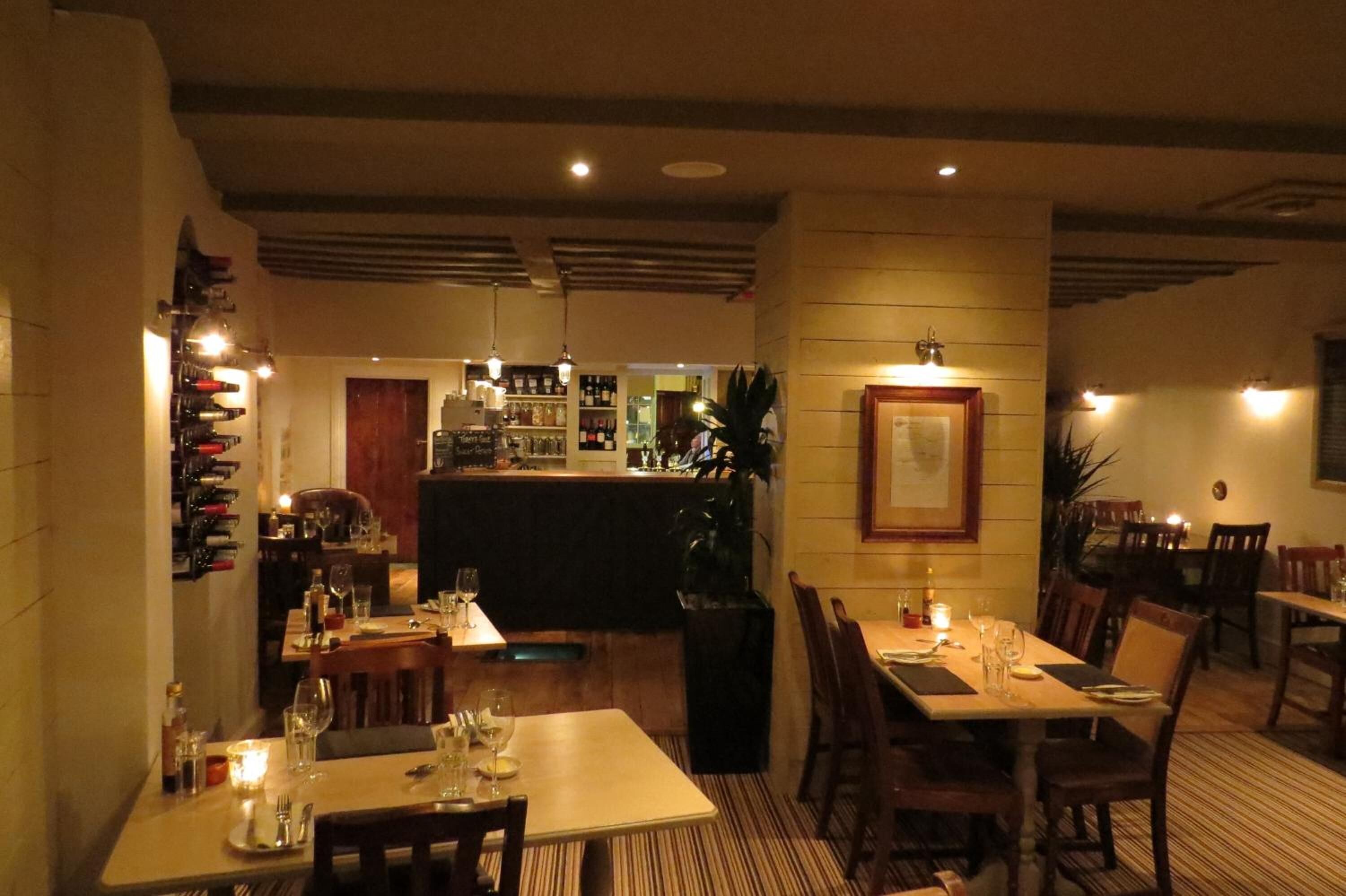 Een restaurant met houten tafels en stoelen, een bar en een wijnrek in de achtergrond.