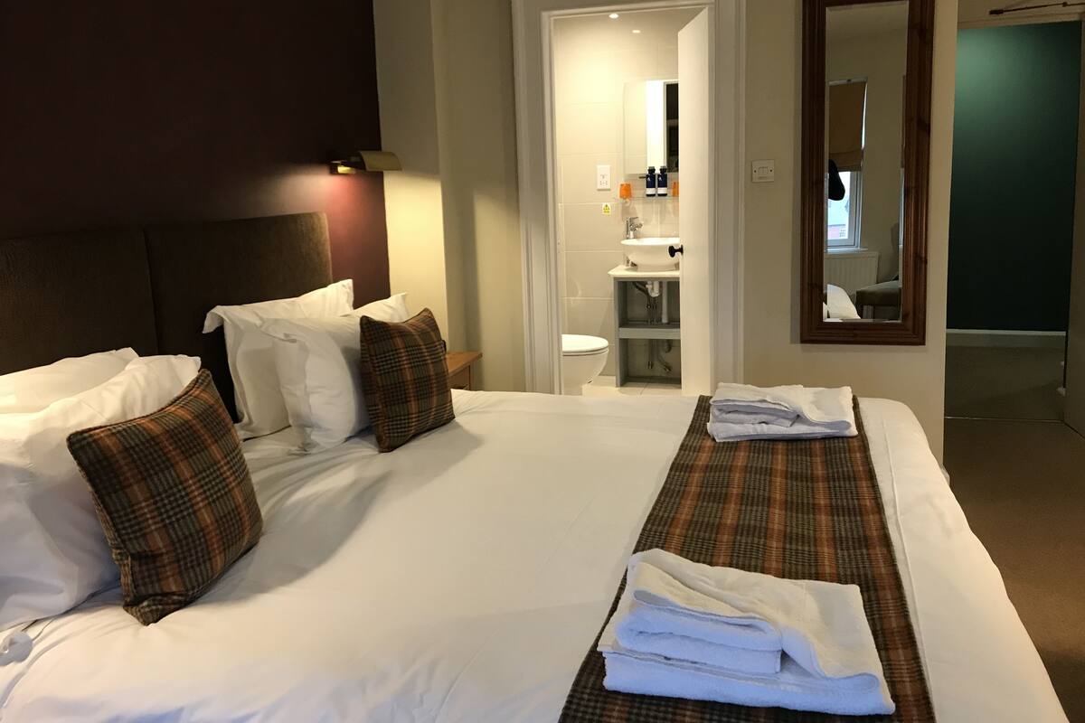 Ein Hotelzimmer mit einem großen Bett, zwei Kissen, einem Nachttisch, einem Spiegel und einem durch eine offene Tür einsehbaren Badezimmer.