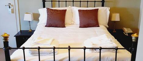 Una cama bien hecha con sábanas blancas y dos almohadas marrones, una mesita de noche con lámpara y un atril con teléfono.