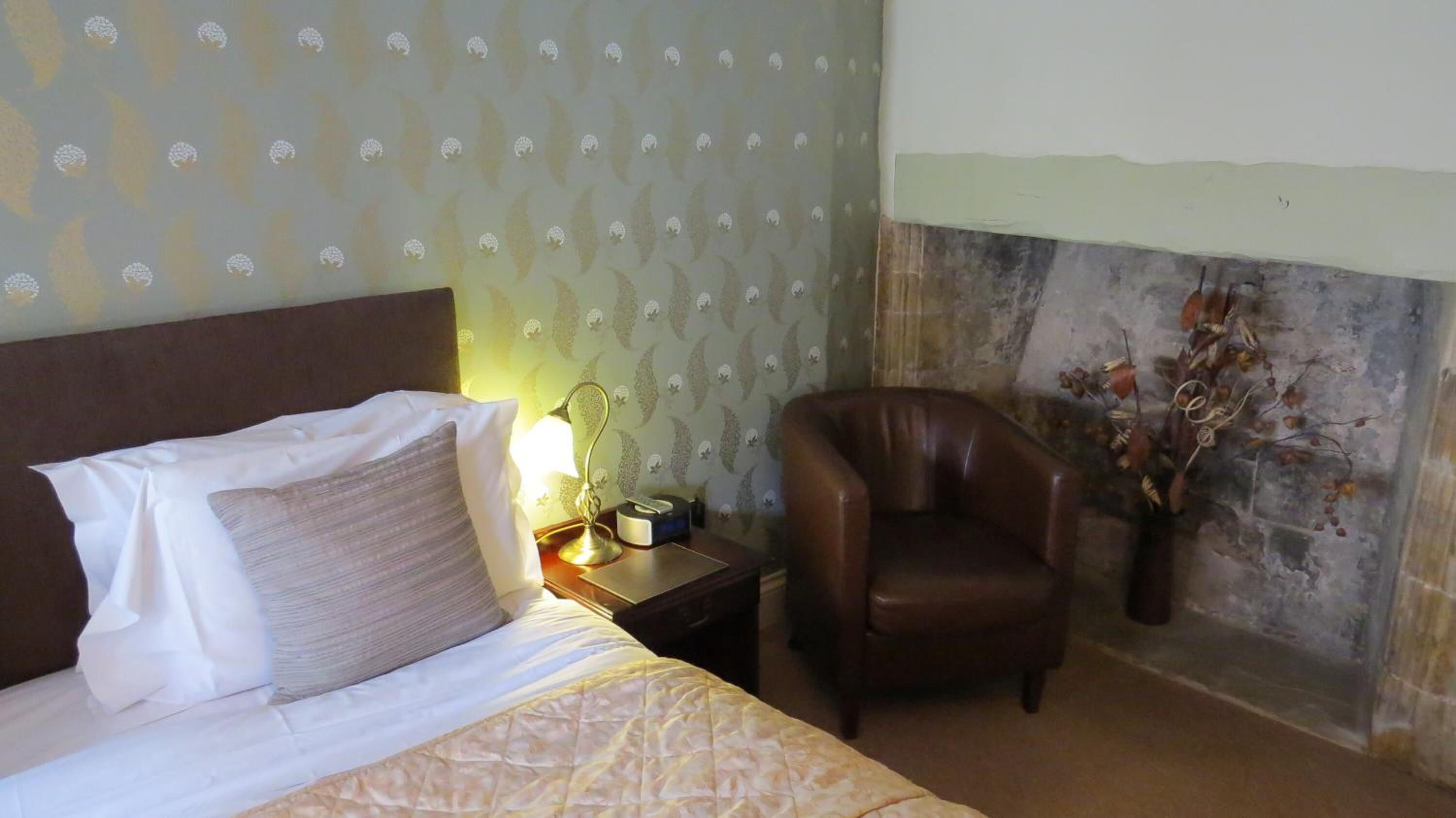 A hotel room with a bed, a nightstand with a lamp, a leather chair, and a decorative wall with a floral arrangement.